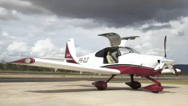 Ein kleines weiß-rotes Propellerflugzeug steht mit offener Cockpittür auf einer Landebahn unter bewölktem Himmel